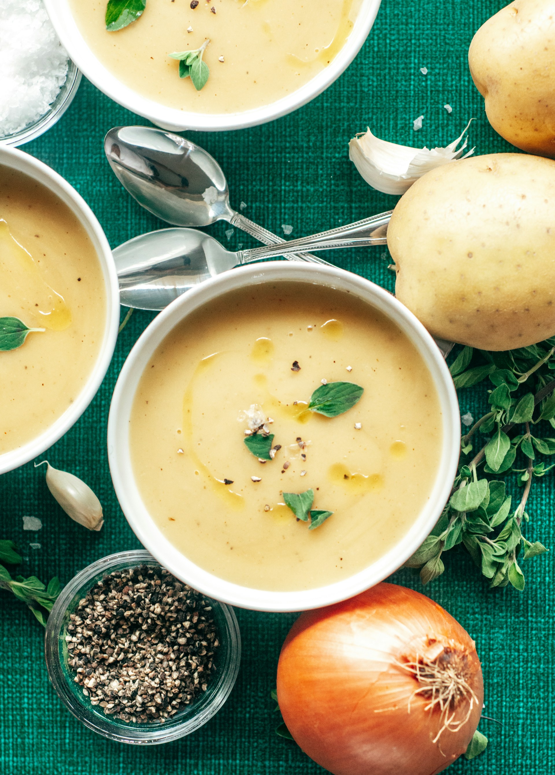 Three white bowls of creamy soup surrounded by potatoes, an onion, garlic, and fresh herbs